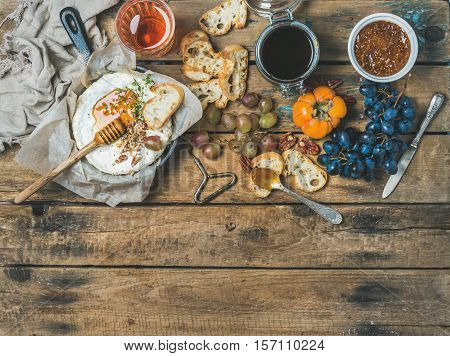 Camembert cheese in small pan, honey, fig jam, persimmon, grapes, pecan nuts, grilled baguette slices and glass of rose wine over rustic wooden background, top view, copy space