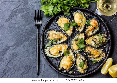 Seafood. Shellfish mussels. Baked mussels with cheese, cilantro and lemon in shells on cast iron black plate. Plate of mussels, cold white wine, lemon and cilantro on black stone background. Top view