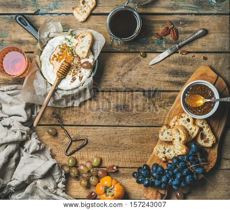 Cheese, fruit and wine set. Camembert in small pan, honey, fig jam, persimmon, grapes, pecan nuts, grilled baguette slices and glass of rose wine over rustic wooden background, top view, copy space