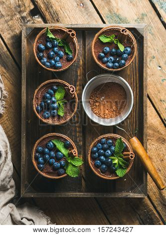 Homemade Tiramisu dessert in glasses with cinnamon sticks, mint leaves, fresh blueberries and sieve with cocoa powder in wooden tray over rustic wooden background, top view, vertical composition