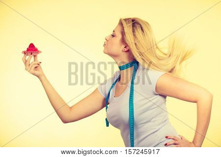 Restrictive diet temptation losing weight concept. Woman holding strawberry sweet cupcake having measure tape around her neck. Studio shot on yellow background