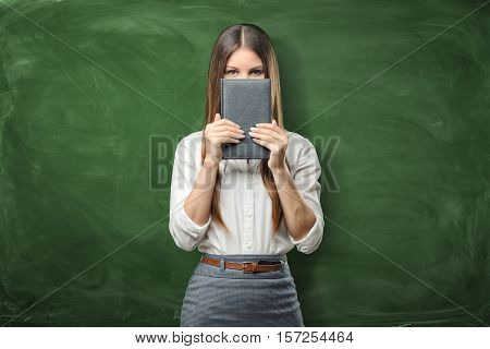 A young businesswoman standing and holding her day planner closed in front of her face on the green chalkboard background. Daily planning. Time-management. Work is done.