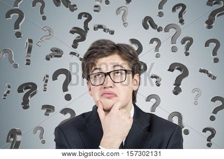 Close up of bespectacled young businessman thinking near a concrte wall with question marks falling. Concept of looking for the answer