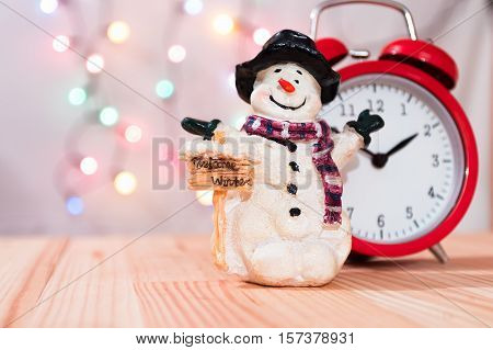 Little snowman figurine with red vintage clock. Natural wooden table. Christmas tree lights bokeh background. Copy space to the left.