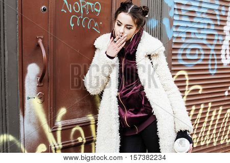 young pretty stylish teenage girl outside at city wall with graffity smoking cigarette at forbidden smoke sighn, modern fashion dressed woman