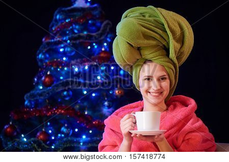 Happy housewife in bathrobe with cup of tea with Christmas tree on background