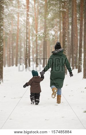 Mother and little toddler boy walking in the winter forest and having fun with snow. Family enjoying winter. Child and woman running outdoors. Winter Christmas and lifestyle concept.