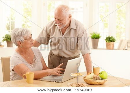 Happy senior couple using laptop computer at home, wife pointing at screen, husband laughing at wife.?