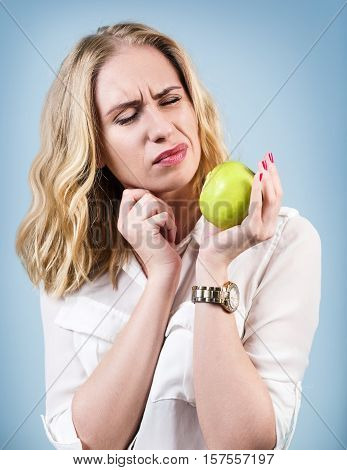 Woman with green apple feels toothpain because of sensitive gums over blue background