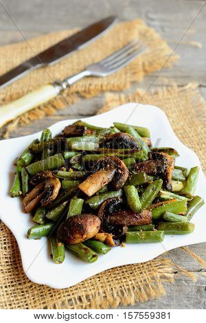 Delicious fried green beans with mushrooms and spices on a plate, fork, knife, burlap on old wooden background. Warm green beans and mushrooms salad. Healthy vegetarian food. Vintage style. Closeup