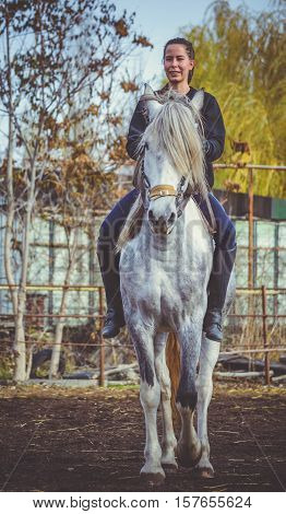 Young beautiful brunette girl rides a horse bareback with hackamore. Warm and sunny autumn day. Portrait of a pretty young woman on the horse, wearing tall boots and gloves.