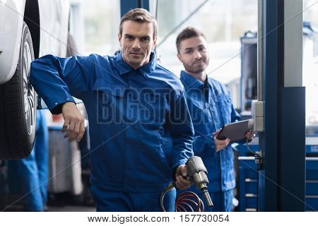 Ucse it smartly.Confident professional car mechanic hoding pneumatic car wrench and going to change the wheel while his colleague standing in the background