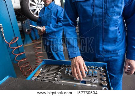 hat to choose. Pleasant professional mechaic standing near set with tools and choosing a correct one while going to repair the car with his colleague