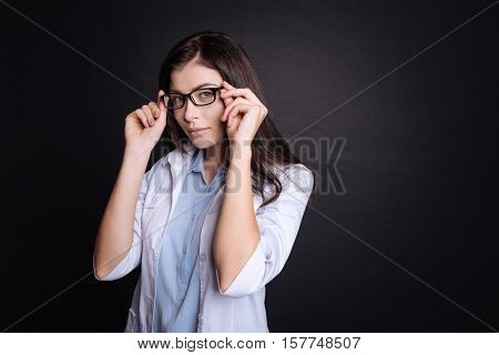 True observer. Pleasant professioanl female doctor touching her glasses and feeling thoughtful while standing isolated on black background