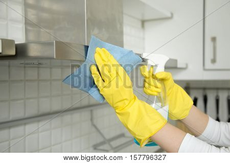 Close up of female hands in rubber protective yellow gloves cleaning the kitchen metal extractor hood with rag and spray bottle detergent. Home, housekeeping concept