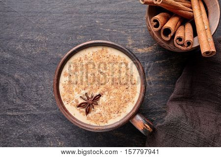 Masala tea chai latte traditional warm Indian sweet milk spiced drink, ginger, cinammon sticks, spices blend organic infusion healthy wellness beverage in rustic clay cup on dark table background