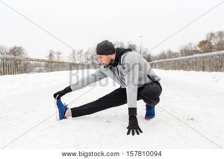 fitness, sport, people, exercising and healthy lifestyle concept - young man stretching leg and warmig up on snow covered winter bridge