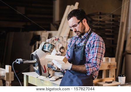 Handsome carpenter in protective glasses standing near his wooden object in the workshop.