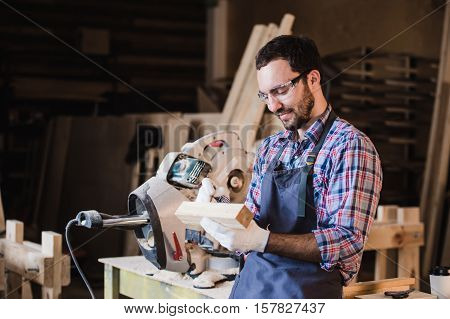 Handsome carpenter in protective glasses is looking at camera and smiling while standing near his wooden object in the workshop.