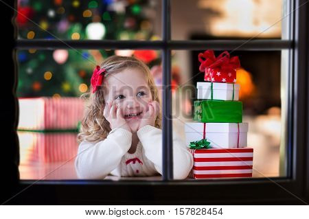 Little girl waiting for Santa at home window on Christmas eve. Kids opening Xmas presents. Child under Christmas tree with gift boxes. View from outside. Winter snowy day at home.