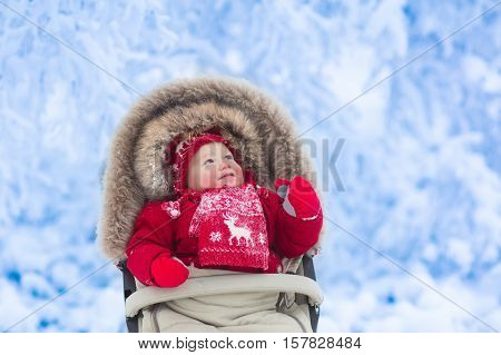 Happy laughing baby in warm red down jacket and knitted Nordic hat and scarf on a walk in a snowy winter park sitting in warm stroller with sheepskin hood catching snow. Child in buggy with foot muff