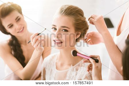 Full of tenderness. Happy delighted young bridesmaids sitting in the white bedroom while helping the bride to get ready and expressing happiness
