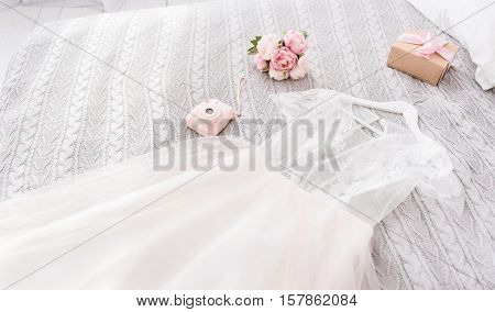 Important elements of the wedding day. White amazing stunning wedding dress and shoes lying on the bed near the pink camera and bouquet in the bedroom