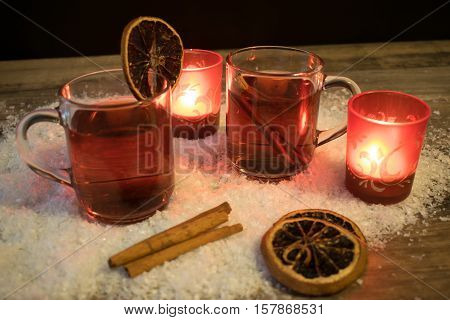 2 glasses with mulled wine in the snow with orange slices cinnamon sticks and 2 red tea light holders with ever a burning tea lights