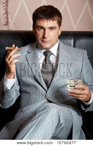Retrato de hombre guapo en traje gris, sentado en el sofá de cuero con whisky y cigarros en casino