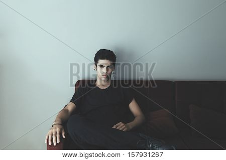 Young handsome brazilian guy with black t-shirt cross earring imposingly sitting on dark red sofa with gray wall behind and looking straight
