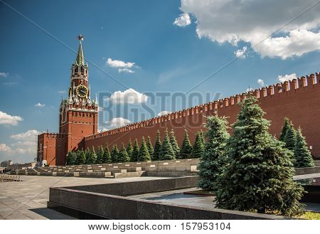 Sunny day at the Red Square with blue sky, white clouds, green fir-trees, the Spasskaya Tower with the Kremlin chimes, the main tower with a through-passage on the eastern wall of the Moscow Kremlin