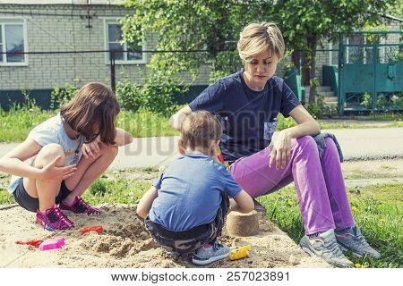 Mom And Children Are Playing On The Playground In The Sand Near The House In Sunny Weather In Summer