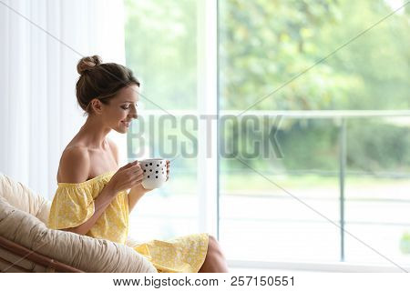 Young Woman With Cup Of Aromatic Coffee Sitting In Papasan Chair Near Window At Home