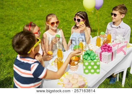 holidays, childhood and celebration concept - happy kids in sunglasses sitting at table on birthday party at summer garden and eating cupcakes