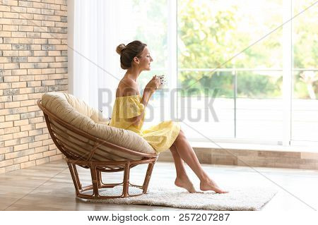 Young Woman With Cup Of Aromatic Coffee Sitting In Papasan Chair Near Window At Home
