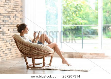 Young Woman With Mobile Phone Sitting In Papasan Chair Near Window At Home