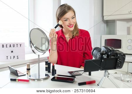 Young Pretty Female Beauty Blogger Sitting At Kitchen Table And Powdering Her Face, Looking At Camer