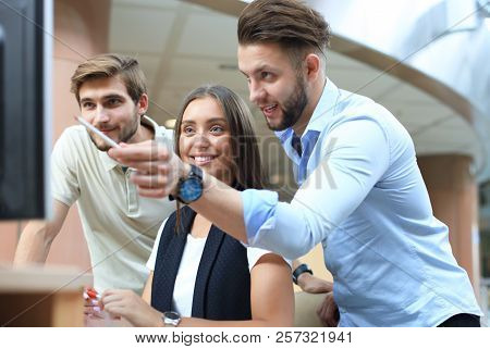 Group Of Young People In Casual Wear Sitting At The Office Desk And Discussing Something While Looki