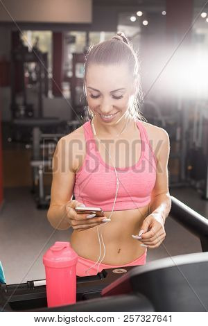 Slim Beautiful Woman With A Ponytale Standing On The Treadmill In The Gym With A Phone Wearing Earph