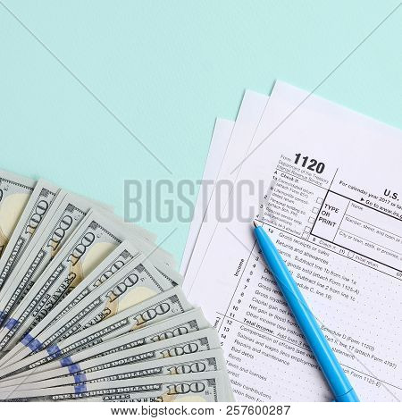1120 Tax Form Lies Near Hundred Dollar Bills And Blue Pen On A Light Blue Background. Us Corporation