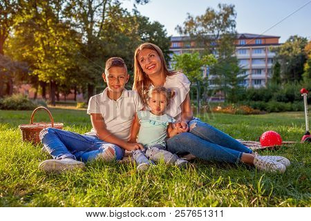 Happy Family Spending Time Outdoors Sittting On Grass In Park. Mom With Two Children Smiling. Family