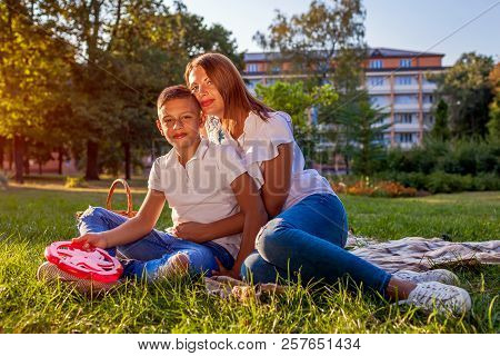 Happy Family Spending Time Outdoors Sittting On Grass In Park. Mother With Her Son Hugging And Smili