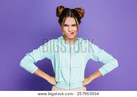 Portrait of angry woman with two buns twisting mouth with furious look isolated over violet background in studio