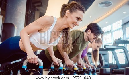 Side view of three young people smiling while doing the kettlebell plank challenge for strong core during group workout at the gym