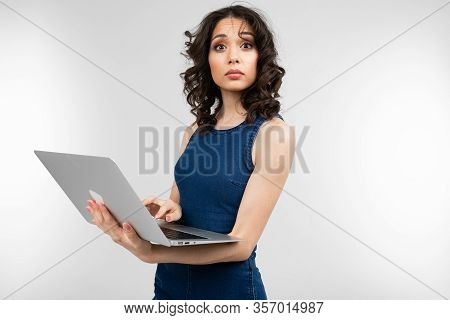 Close-up Of A Surprised Girl In A Blue Dress Holding A Laptop In Her Hands On A White Studio Backgro