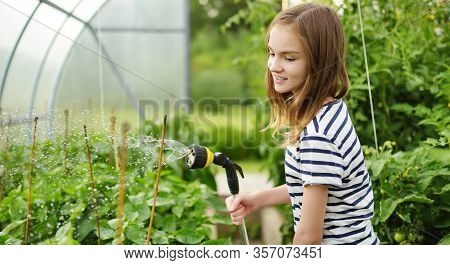 Adorable Young Girl Watering Variuos Vegetables In A Greenhouse On Sunny Summer Day. Gardening Activ