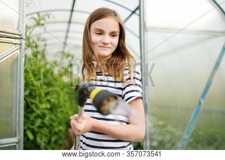 Adorable Young Girl Watering Variuos Vegetables In A Greenhouse On Sunny Summer Day. Gardening Activ