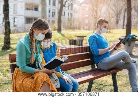 People sitting on park bench in the sun practicing social distancing in corona crisis wearing masks