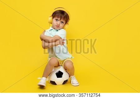 Full Lengh Photo Of Upset Little Boy Sitting On Soccer Ball Wearing Headphone Over Yellow Background
