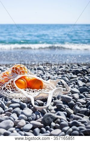 White Cotton Eco String Bag With Oranges On A Rocky Beach. Sea View With A Blue Sky. Zero And Eco Wa